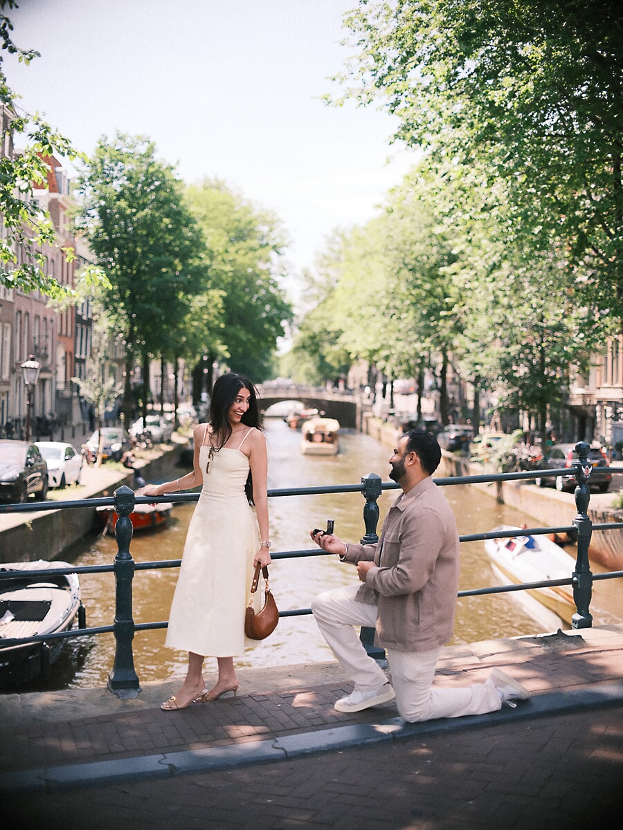 Man kneeling with an open ring box on a canal bridge as his partner turns to him, an Amsterdam proposal photoshoot