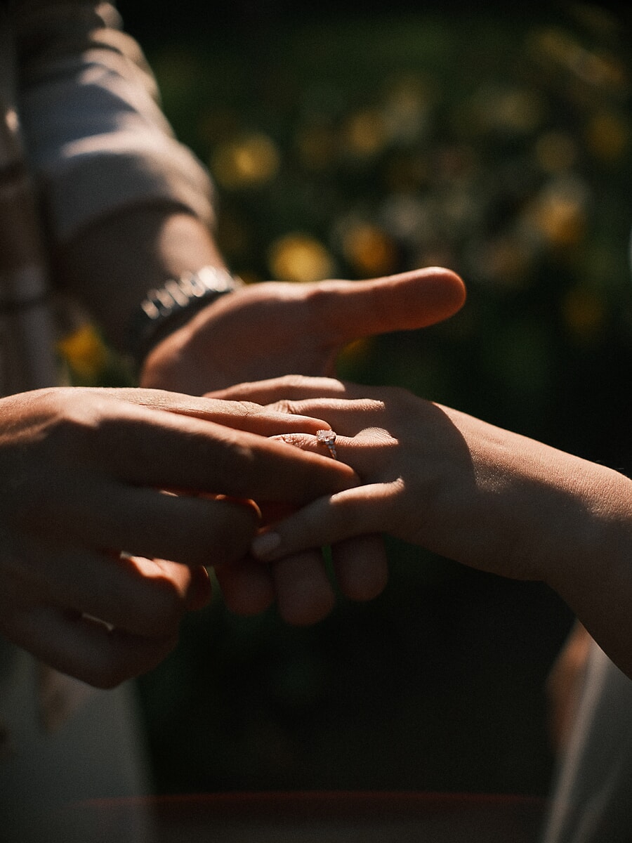 Two hands meeting as the proposal ring is placed, sunlit flowers blurred behind