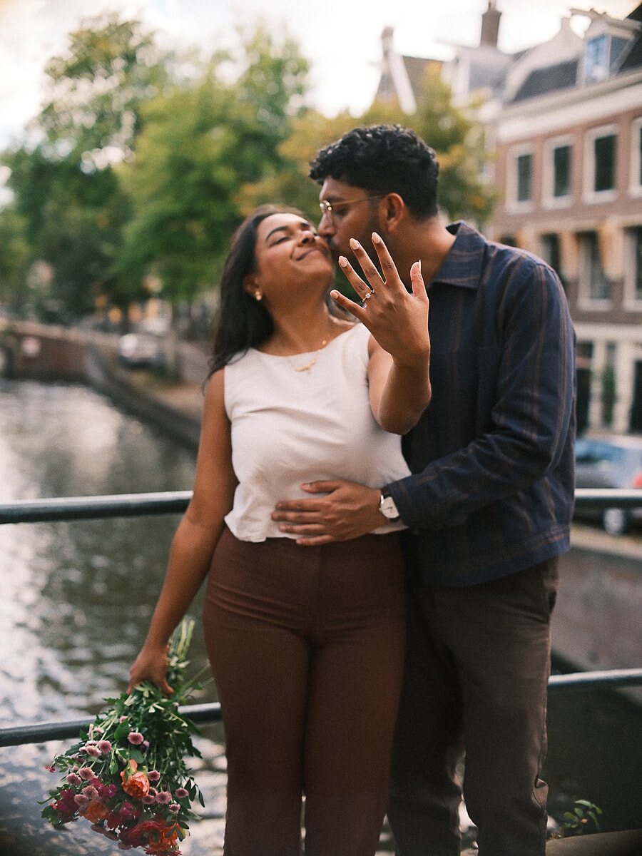 Man kissing his fiancée's temple beside an Amsterdam canal as she raises her hand to show the engagement ring