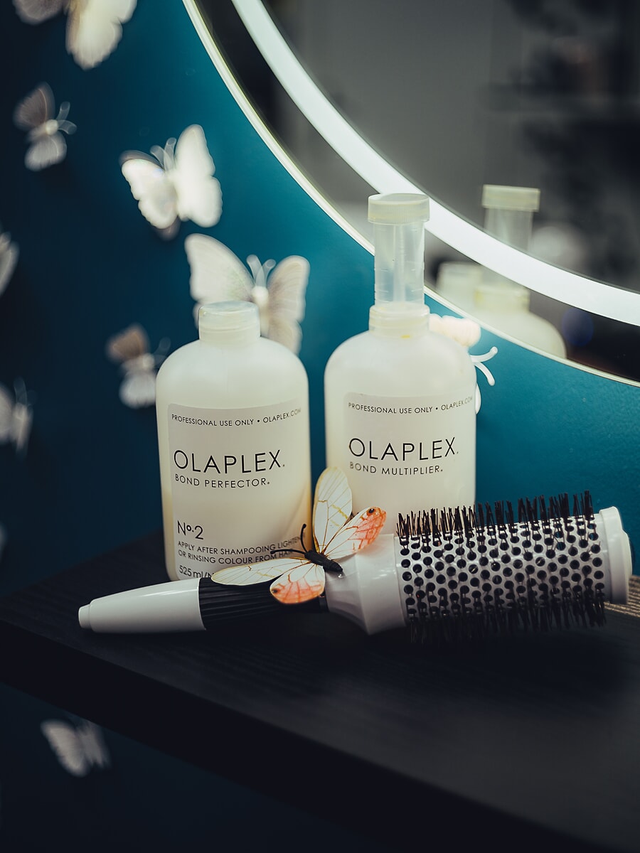 Two salon treatment bottles and a round brush on a black shelf against a teal wall with paper butterflies