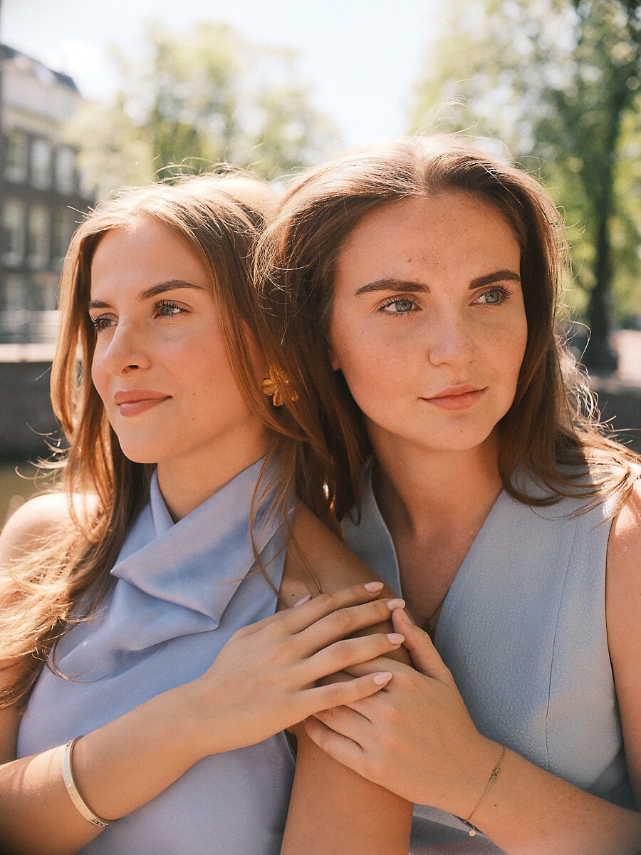 Two sisters in pale blue standing close together in warm backlight, one resting against the other's shoulder, hands clasped