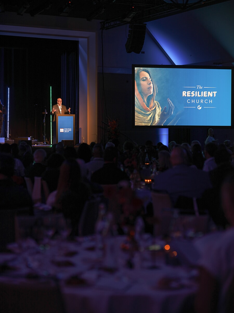 Speaker at a lectern seen over the heads of a seated dinner audience, the title slide lit blue behind him