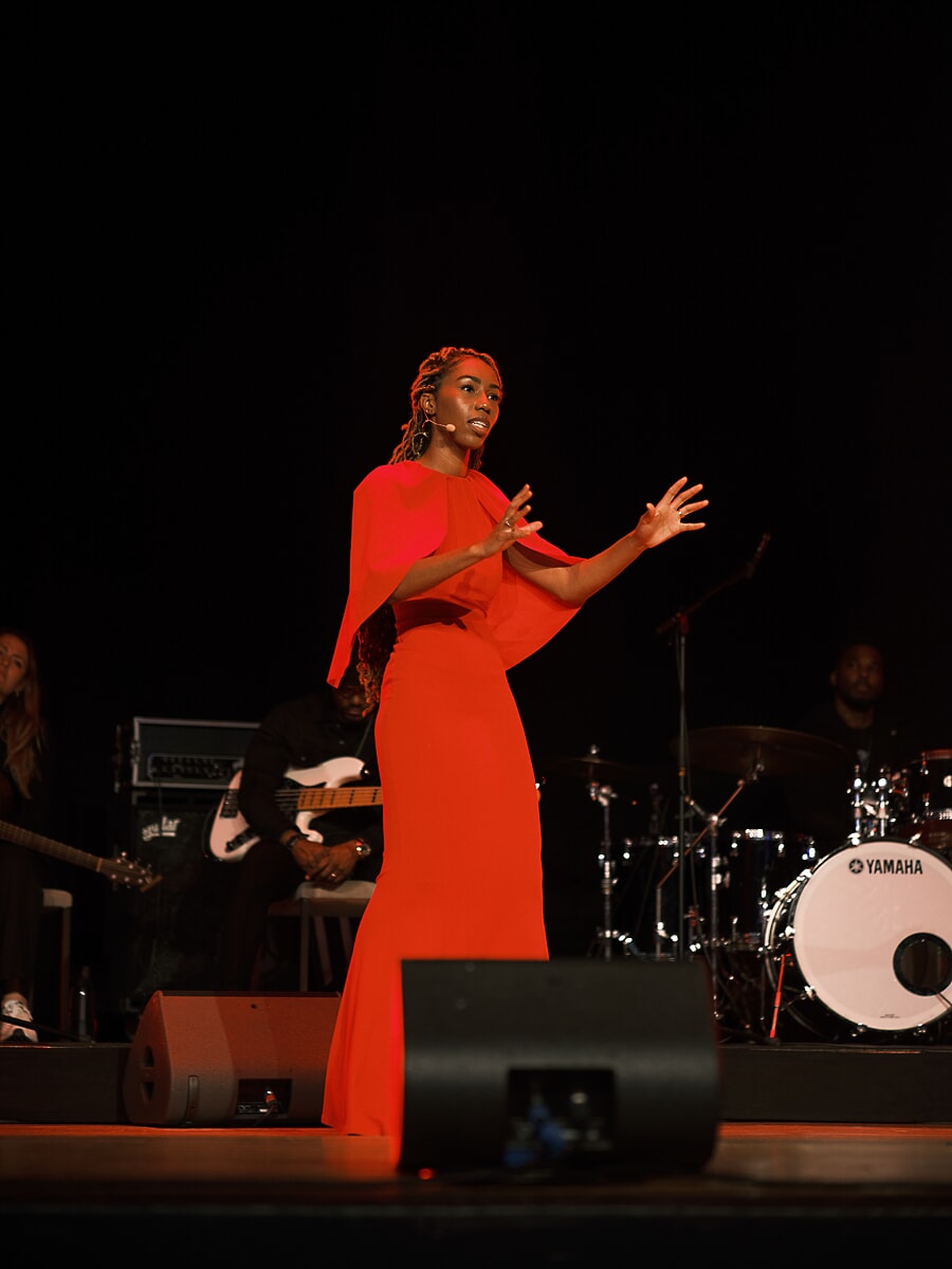 Speaker in a red gown on a darkened stage with a headset microphone, the band's drum kit behind her