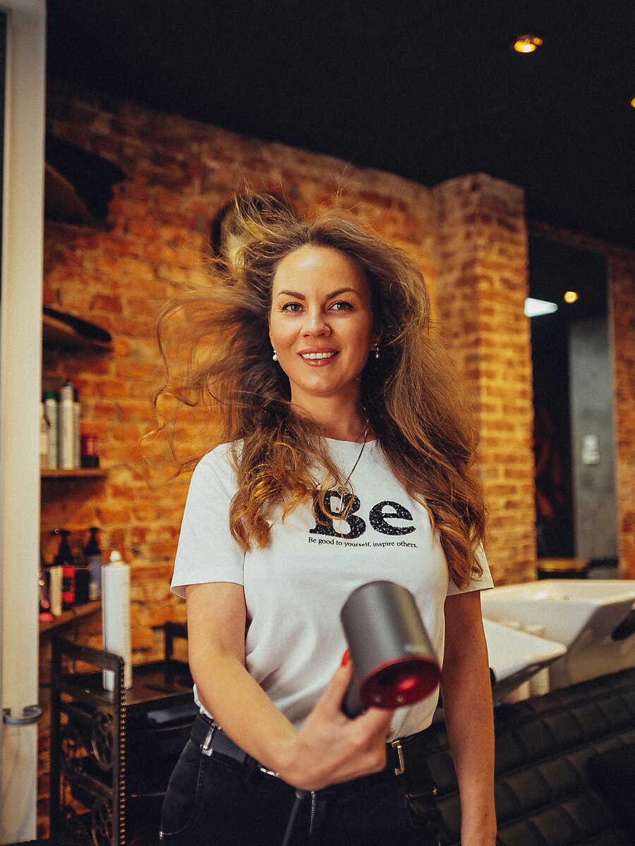 Stylist holding a hairdryer as the air lifts her hair, exposed brick and product shelves behind her, product photography for an Amsterdam business