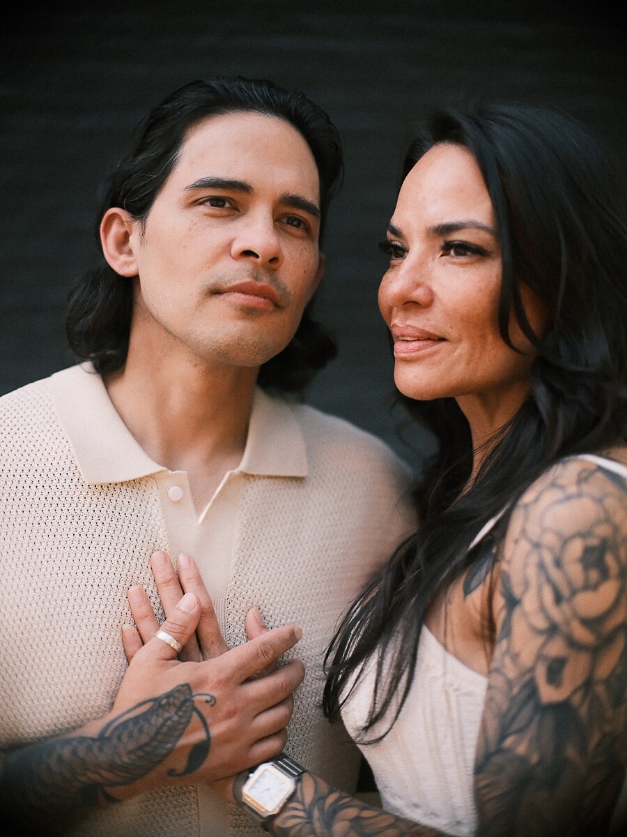 Close portrait of a tattooed couple against a dark wall, their hands clasped over his chest, she rests against his shoulder