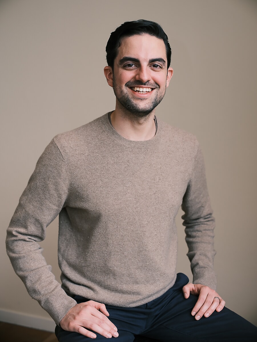 Colleague in a light beige jumper seated with his hands on his knees against a warm neutral wall, a team portrait for an Amsterdam business