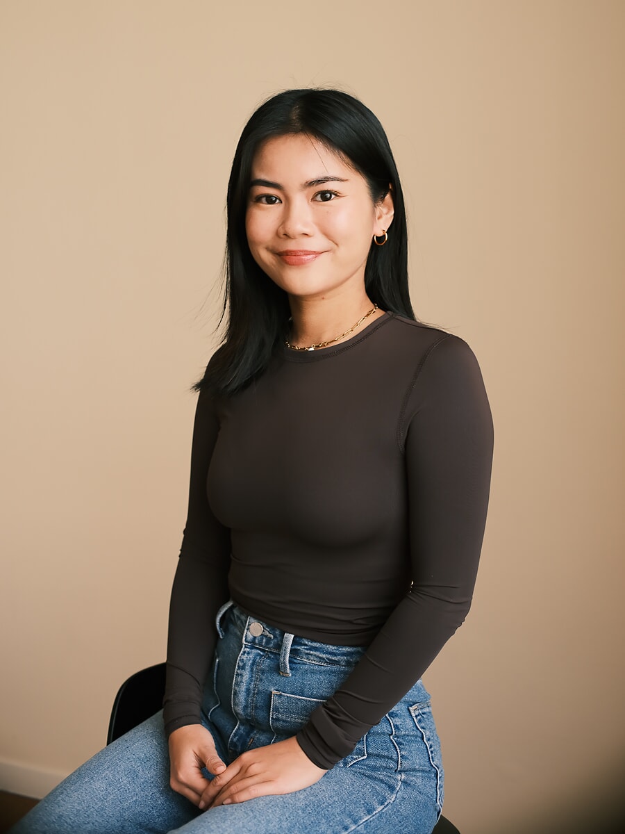 Colleague in a brown long-sleeve top and jeans seated with her hands in her lap against a plain wall