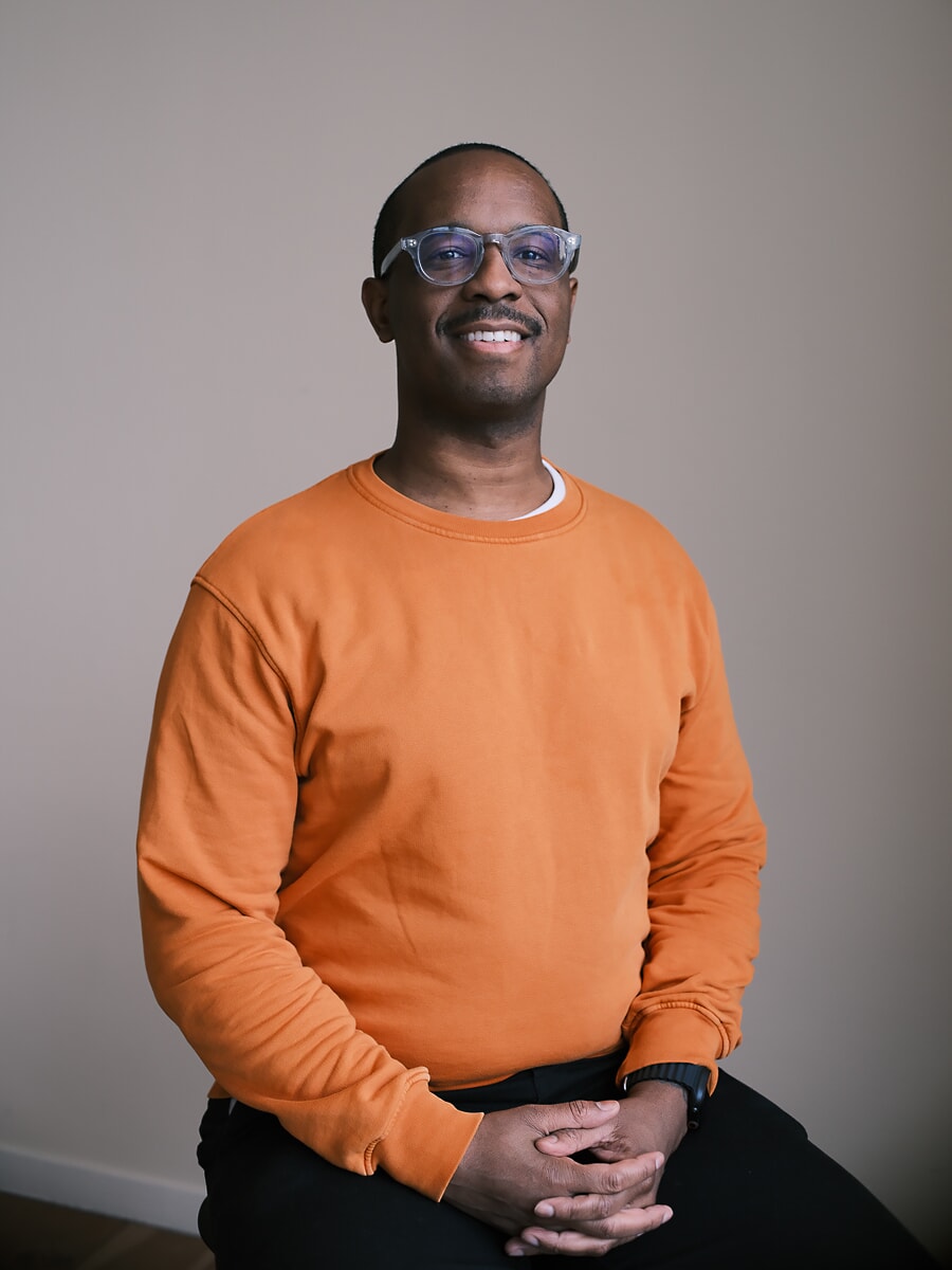 Colleague in an orange sweatshirt and clear-framed glasses, seated with his hands clasped, smiling to camera