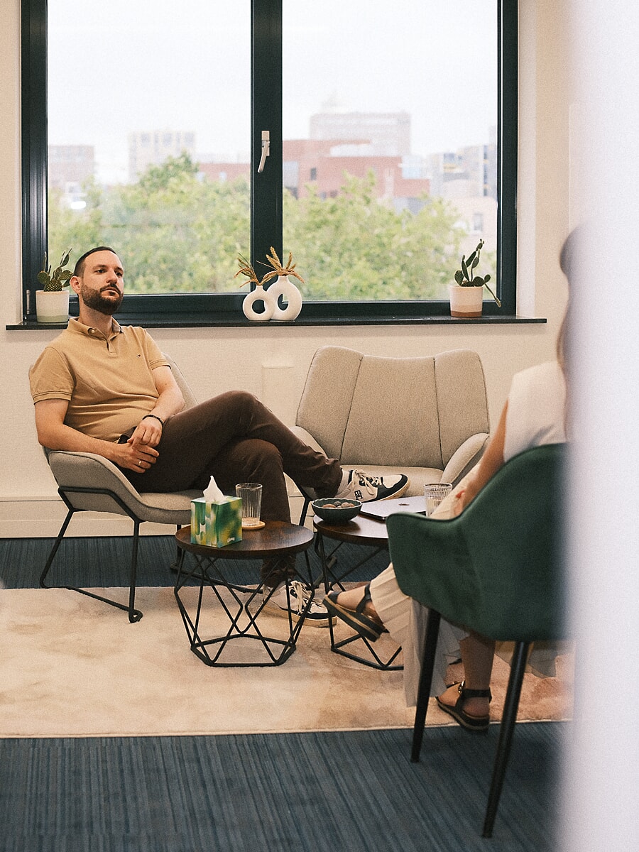 Therapist in a tan polo listening from a low chair by a wide window, rooftops beyond, a branding shoot for an Amsterdam business
