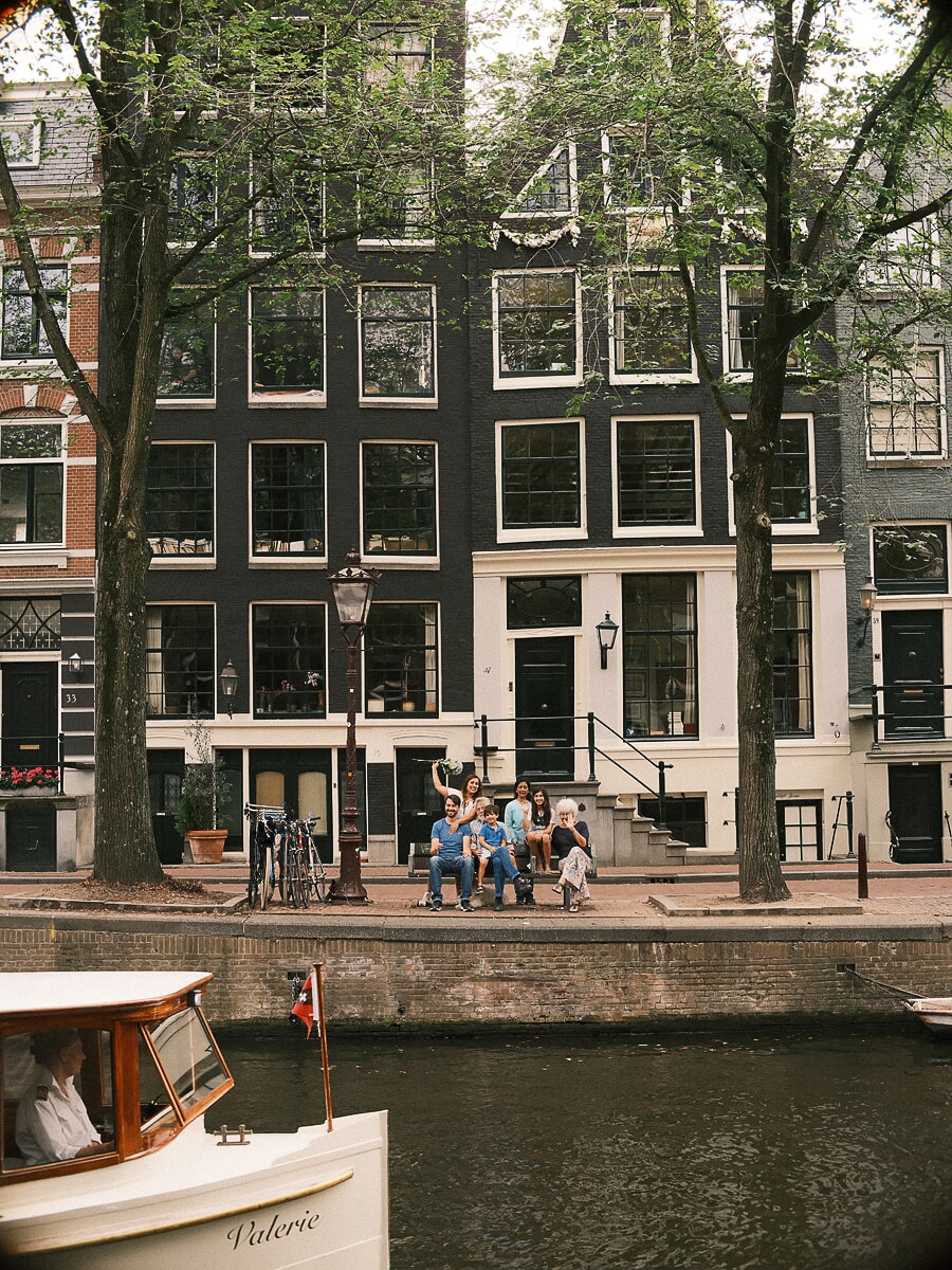 Three generations of one family gathered on the steps of a canal house in Amsterdam, seen from across the water as a saloon boat passes