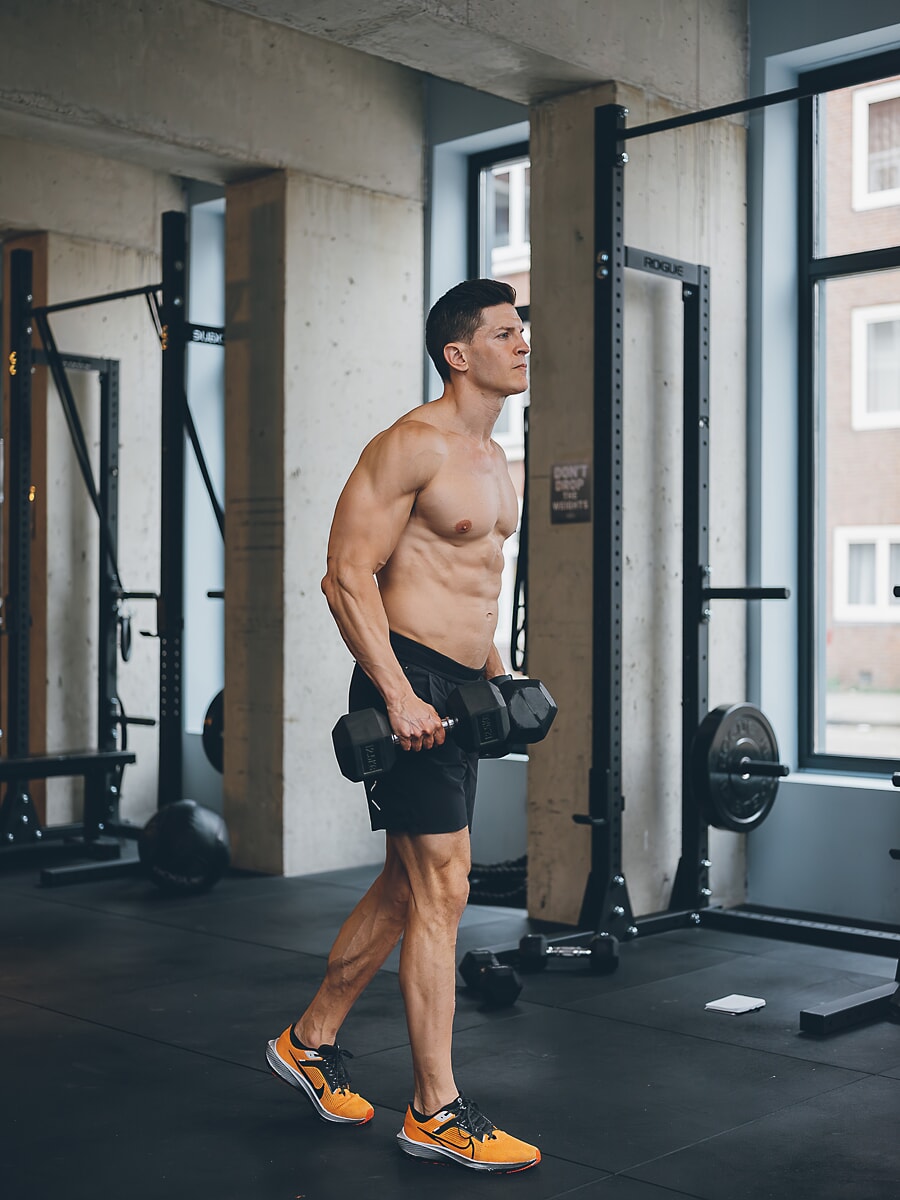 Trainer carrying two dumbbells across a concrete gym floor, squat racks and tall windows behind him