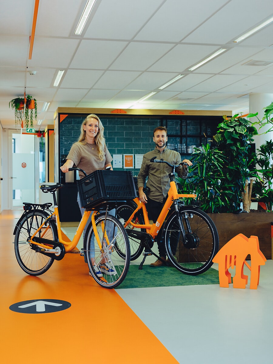 Two staff with a pair of orange e-bikes in a bright office, orange floor markings behind them, a business photoshoot in Amsterdam