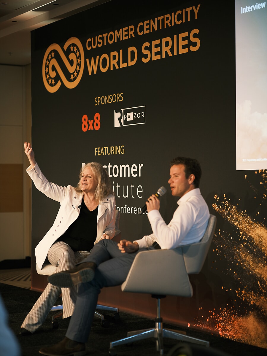 Two speakers in conversation on stage at a conference in Amsterdam, one of them holding a microphone