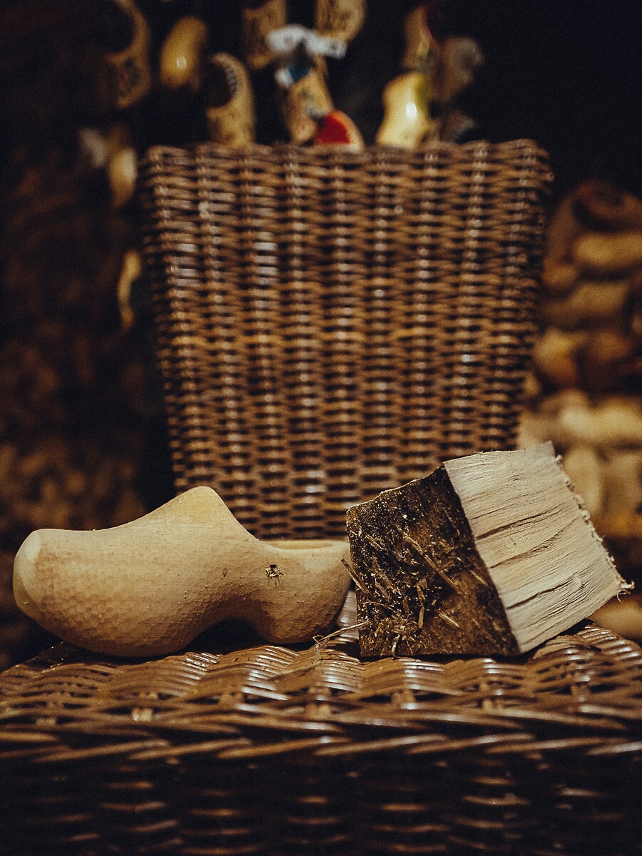 Unfinished wooden clog resting beside a split log on a wicker basket, painted clogs blurred behind