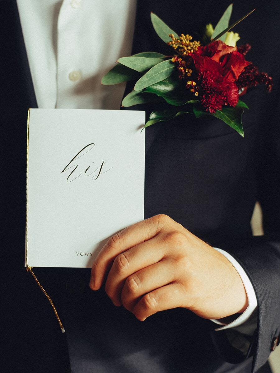 Wedding Ceremony SP10 A 'his' vow book held in the groom's hand with a deep red boutonnière on his lapel, an intimate ceremony detail from the wedding photo session