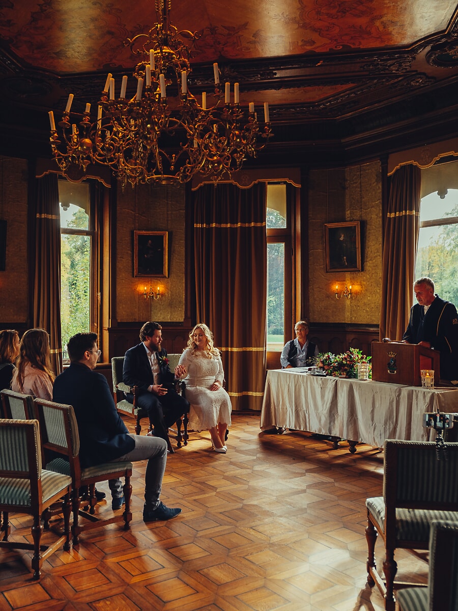 Wedding Ceremony SP13 Civil ceremony in a chandelier-lit hall, the couple seated before the registrar, documented by a wedding photographer in Amsterdam