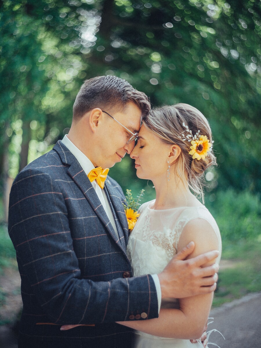 Wedding Ceremony SP14 Groom in a checked suit resting his forehead against his bride's on a leafy green path, a sunflower in her hair, a tender wedding portrait