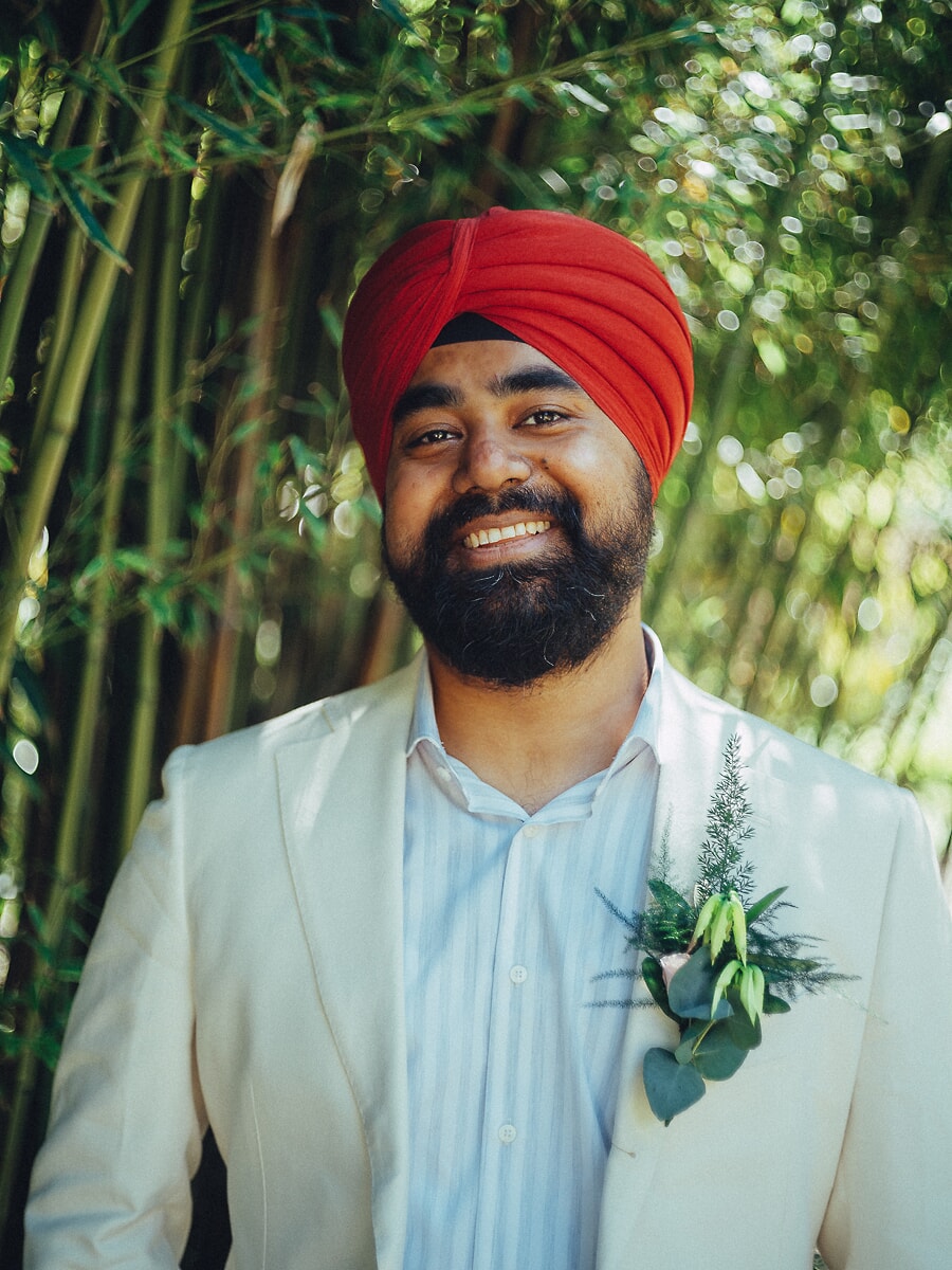 Wedding Ceremony SP21 Smiling groom in a red turban and cream suit with a green boutonnière before lush bamboo, a joyful wedding portrait