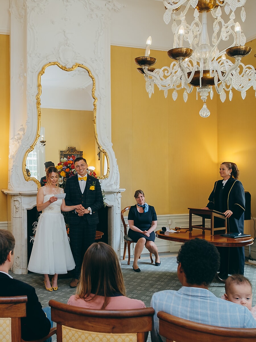 Wedding Ceremony SP23 Vows in hand, the bride stands before the registrar in a gilded city hall room, guests watching in a candid civil ceremony photo