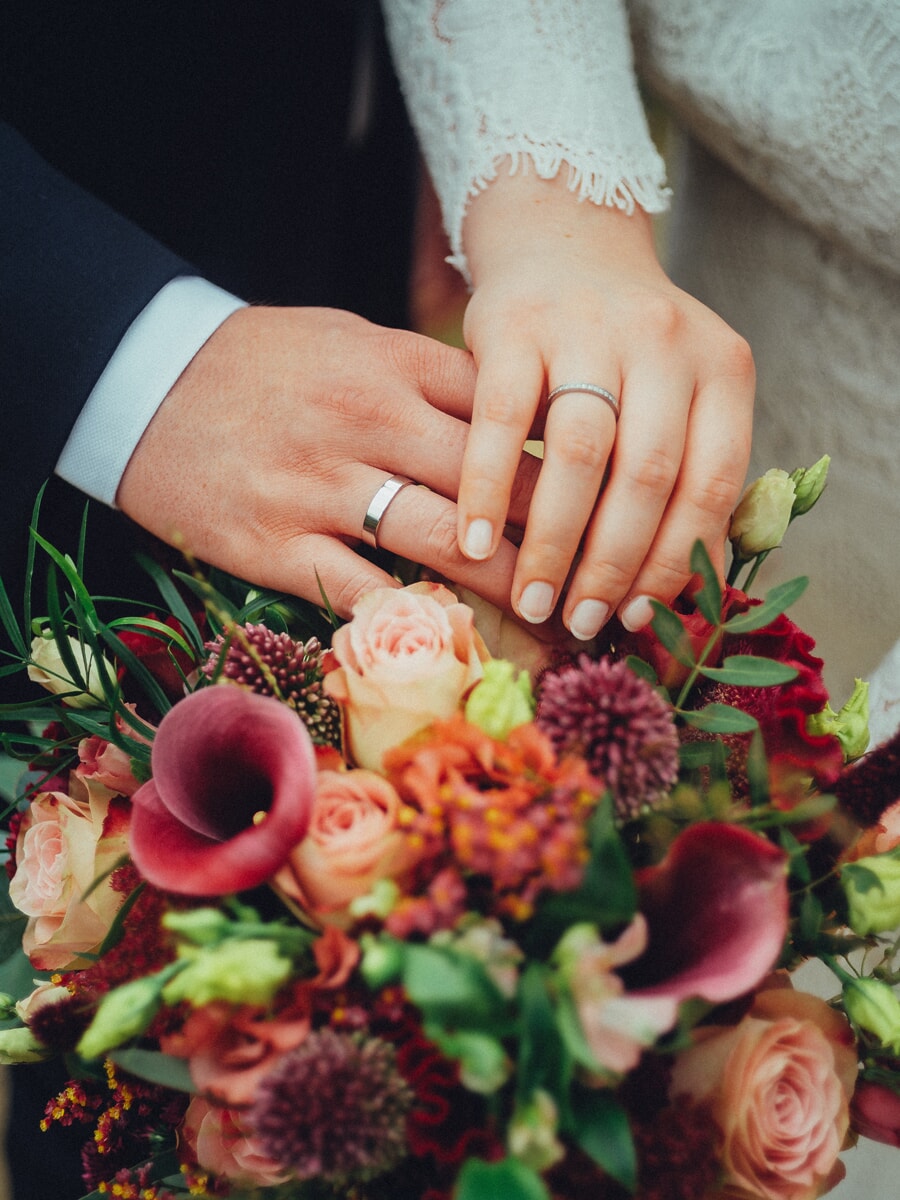 Wedding Ceremony SP26 Newlyweds resting their ringed hands on an autumn bouquet of roses and calla lilies, an intimate wedding photo