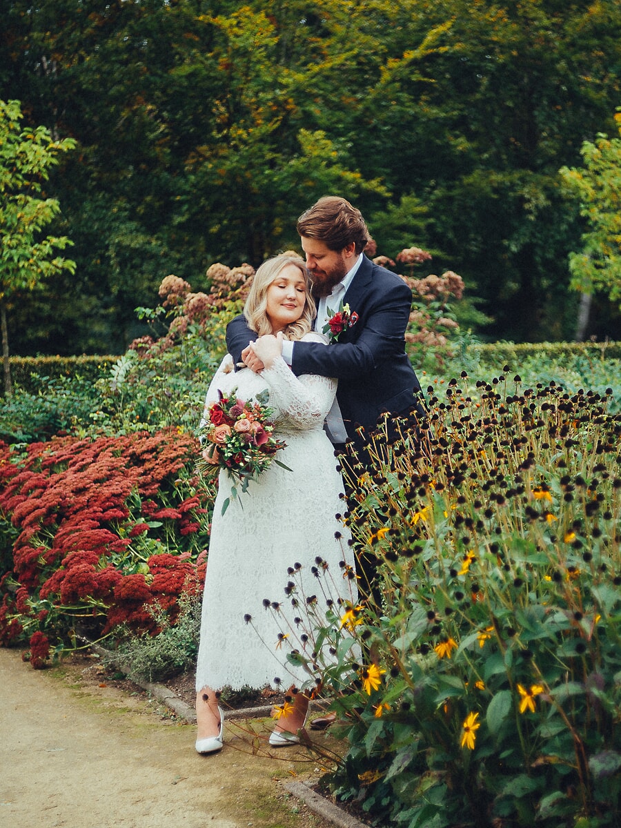 Wedding Ceremony SP27 Groom embraces his bride from behind in an autumn garden of red blooms, her bouquet in hand, by a wedding photographer in Amsterdam