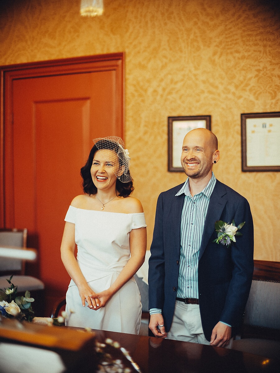 Wedding Ceremony SP28 Laughing bride in an off-shoulder gown and birdcage veil beside her groom inside an Amsterdam city hall, captured by a wedding photographer in Amsterdam