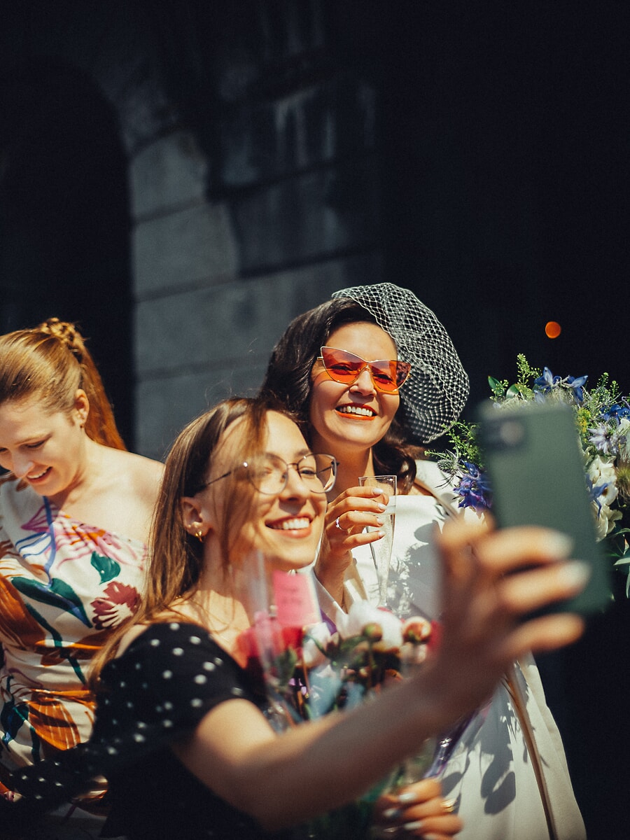 Wedding Ceremony SP29 In a birdcage veil and cat-eye sunglasses, the bride takes a selfie with laughing guests, a candid wedding photo