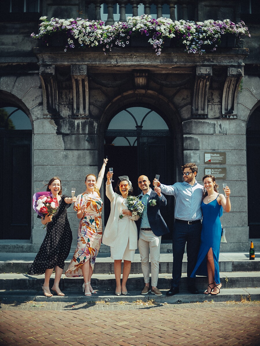 Wedding Ceremony SP30 A wedding photographer in Amsterdam catches newlyweds and guests raising glasses on the steps of a flower-topped city hall