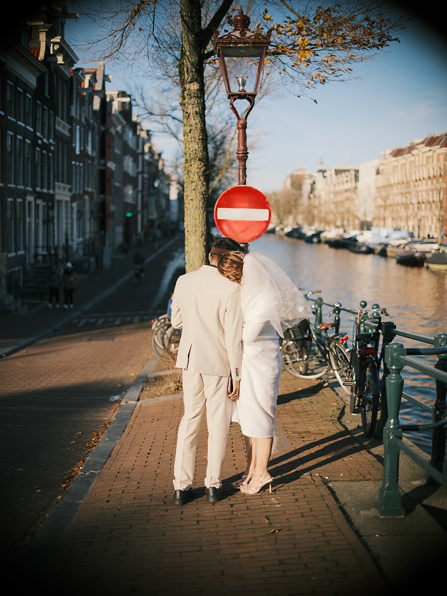 Wedding Ceremony SP4 Couple in cream wedding attire walking hand in hand down a sunlit Amsterdam canal street past parked bicycles, captured by a wedding photographer in Amsterdam