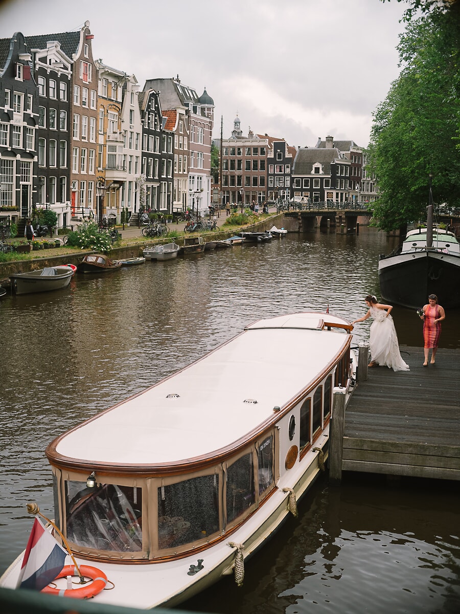 Wedding Ceremony SP7 White salon boat moored along a canal lined with historic houses as the bride steps aboard, a wide frame of Amsterdam wedding photography