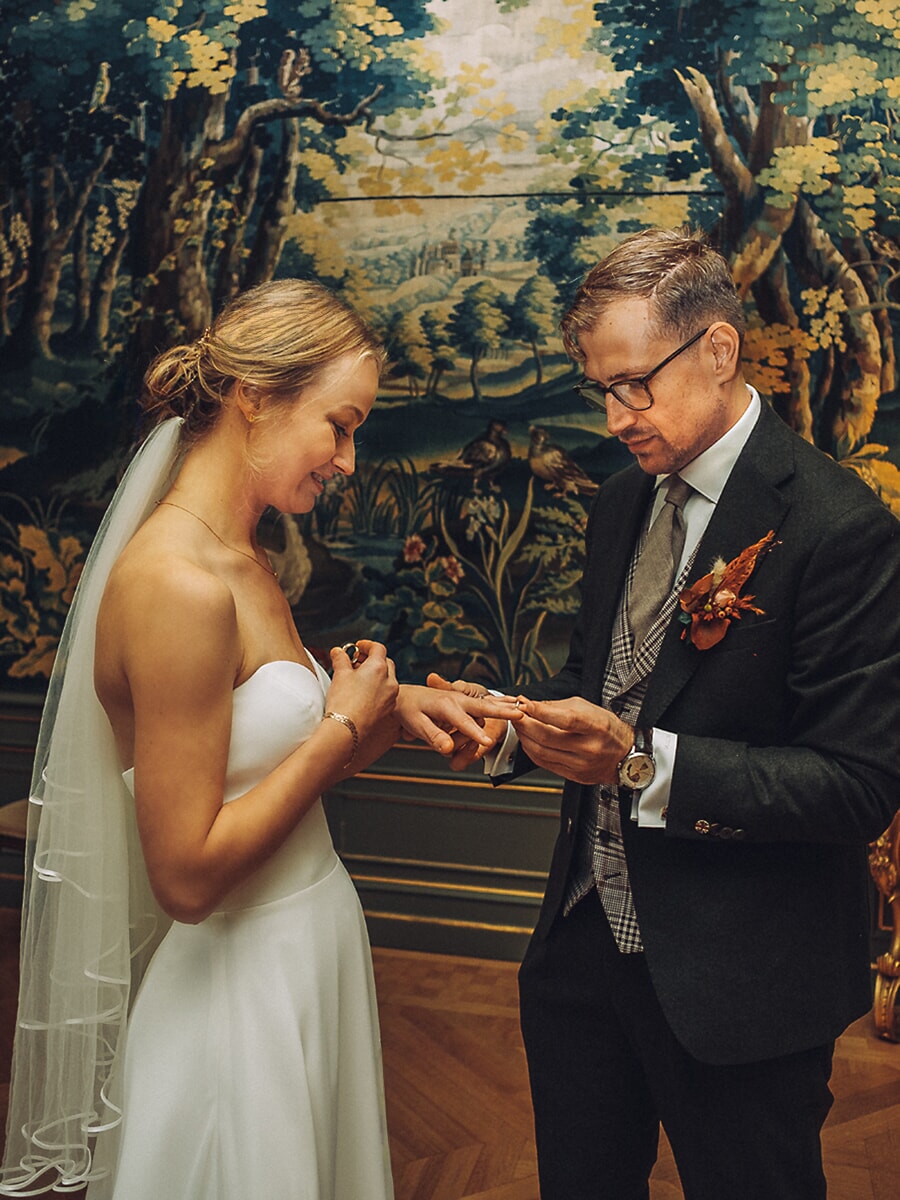 Wedding Ceremony SP34 Bride slides the ring onto her groom's finger before a woodland tapestry in a parquet-floored hall, a tender civil ceremony moment