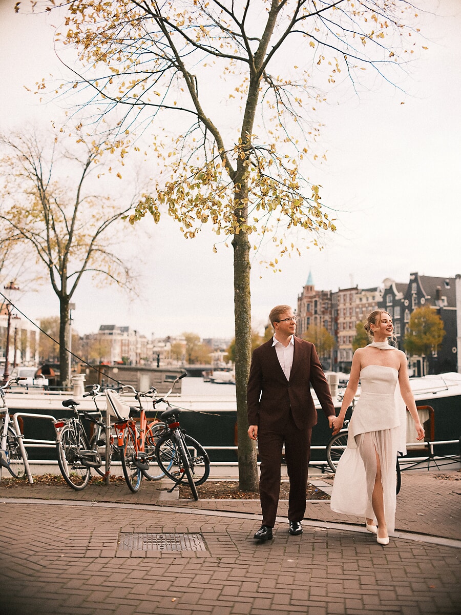 Wedding Bridal SP10 Groom in brown corduroy and bride in a pleated ivory skirt walk under an autumn tree along the Amstel, a relaxed newlywed photo