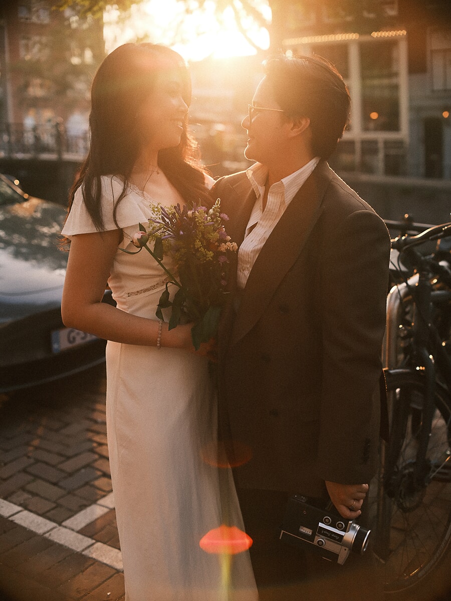 Wedding Bridal SP12 Golden hour light falls on the couple beside a canal as they hold a wildflower bouquet and an old film camera, a romantic elopement frame