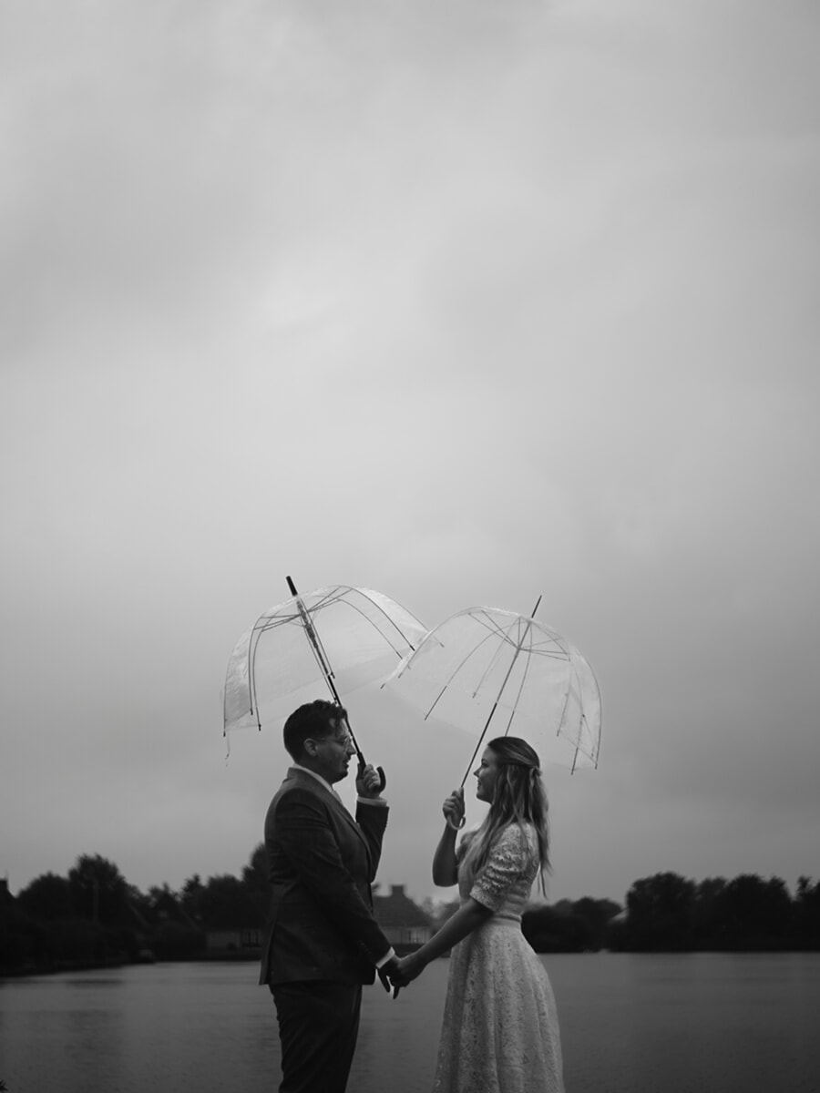 Wedding Bridal SP18 Black and white portrait of a couple holding hands under two clear umbrellas beside misty water, by a wedding photographer in Amsterdam