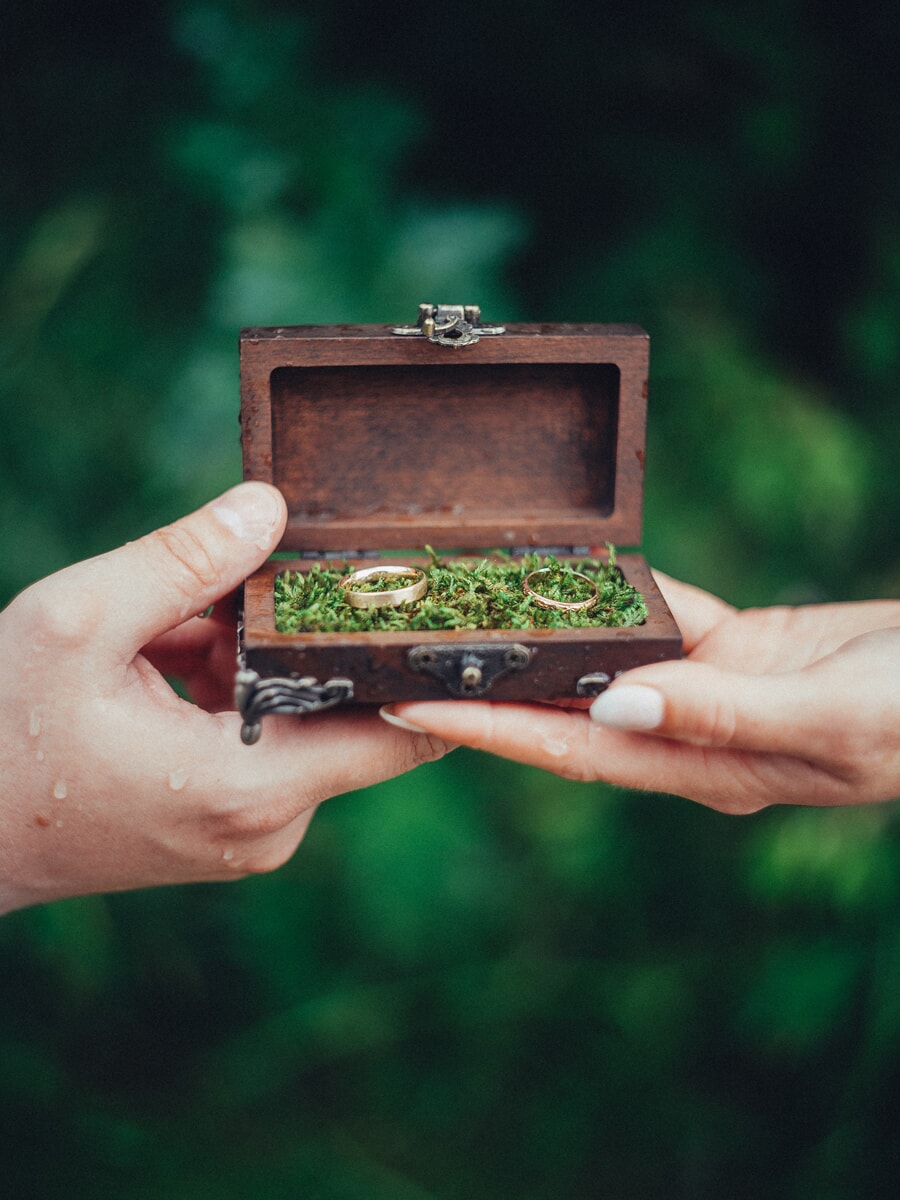Wedding Bridal SP19 Two hands hold open a small wooden box lined with moss and two gold rings against deep green foliage, an artistic elopement detail