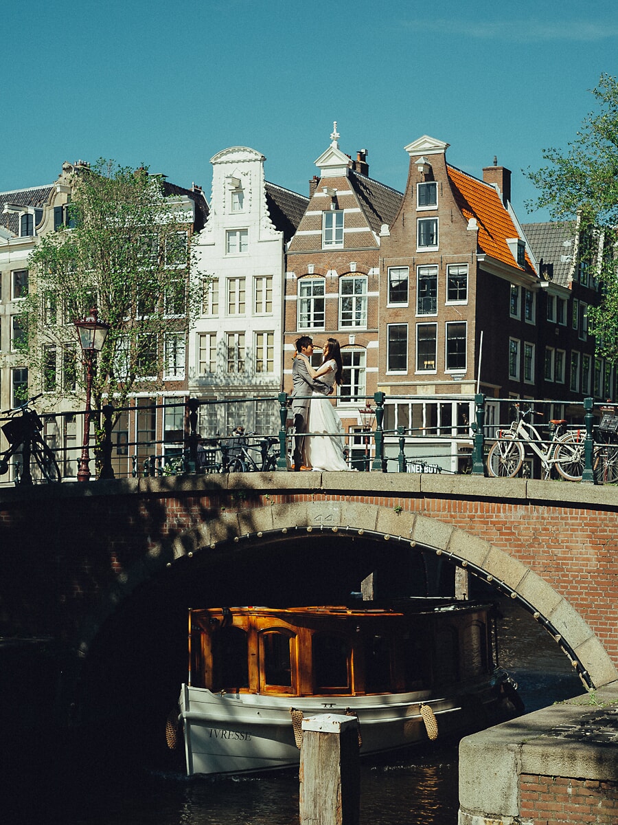 Wedding Bridal SP20 Newlyweds embrace on a canal bridge before gabled facades, a saloon boat moored below, by a wedding photographer in Amsterdam