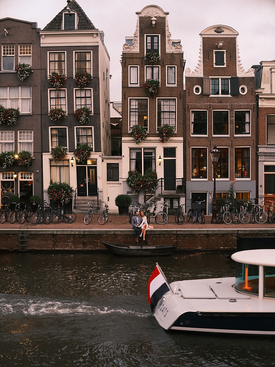 Wedding Bridal SP3 Below flower-decked gables the couple sit at the canal's edge as a boat passes, a cinematic frame by a wedding photographer in Amsterdam