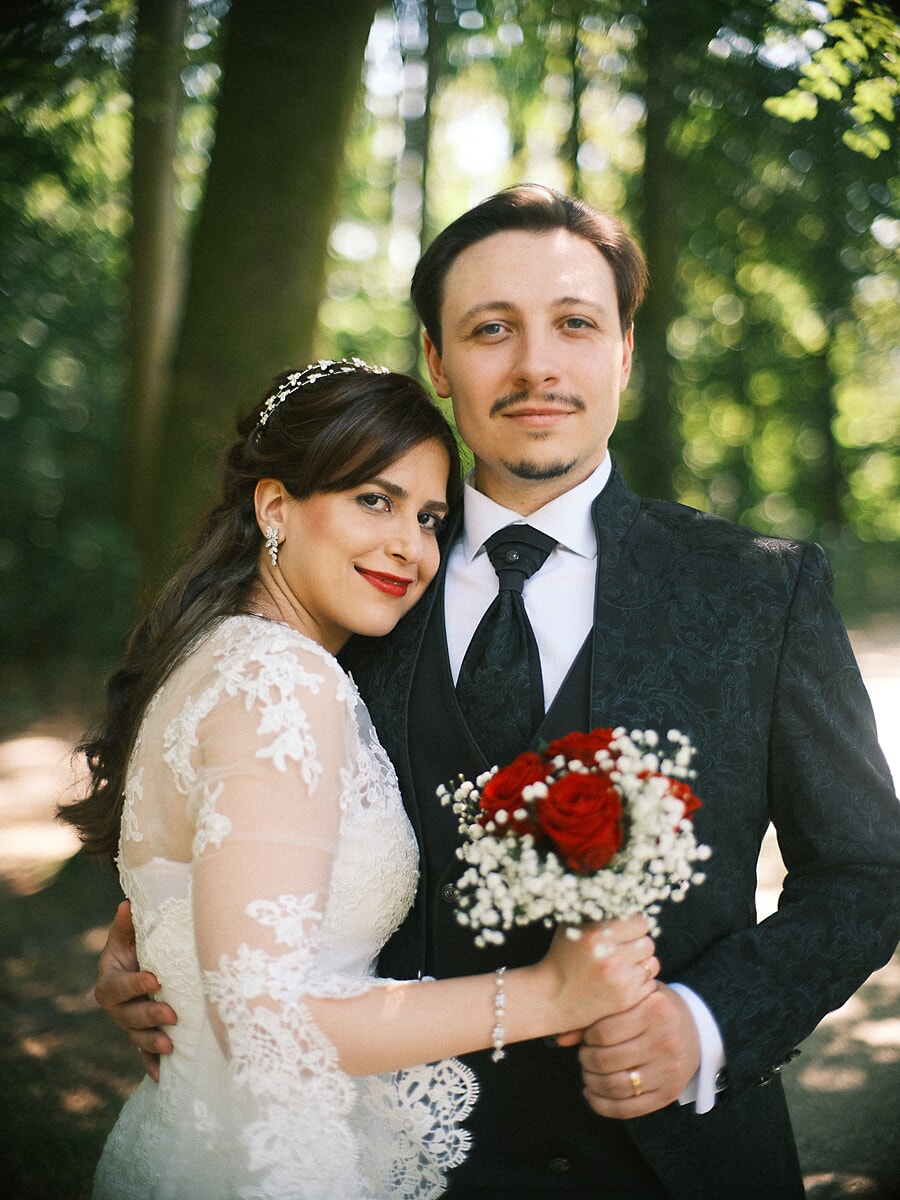 Wedding Bridal SP4 Groom in a patterned black suit holds his bride in lace sleeves and a pearl headband in a sunlit forest, an elegant elopement portrait