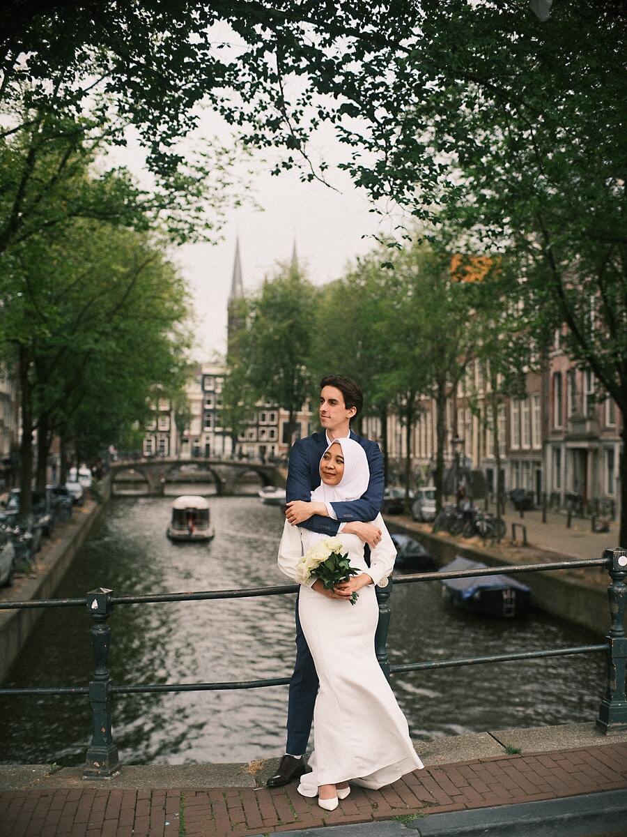 Wedding Bridal SP5 In an ivory gown and hijab, the bride leans into her groom's arms on a canal bridge, shot by a wedding photographer in Amsterdam
