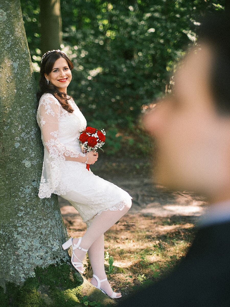 Wedding Bridal SP6 Bride in a lace dress leans against a mossy tree with red roses, her groom blurred in the foreground, a romantic elopement photo