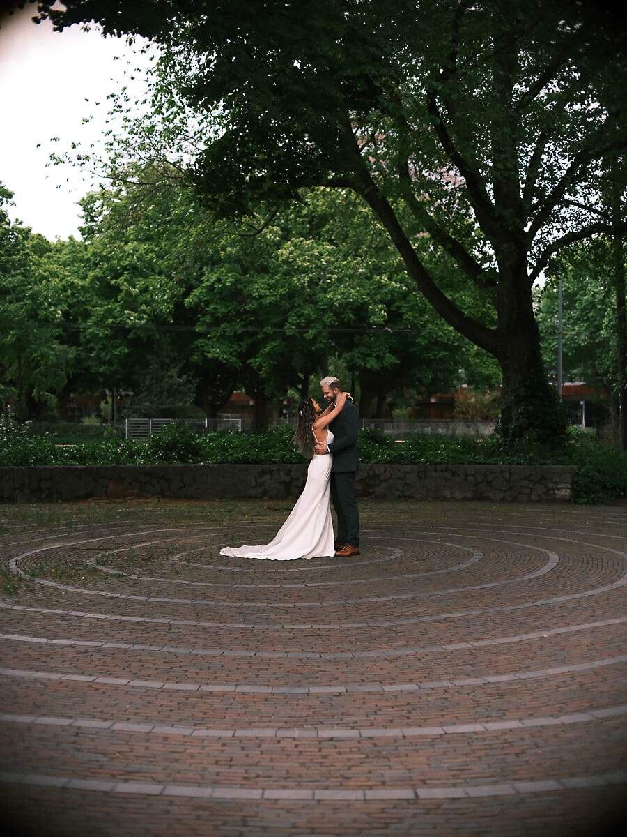 Wedding Bridal SP9 Couple embrace at the centre of a circular brick labyrinth, the bride's train fanned across the stone, an atmospheric elopement portrait