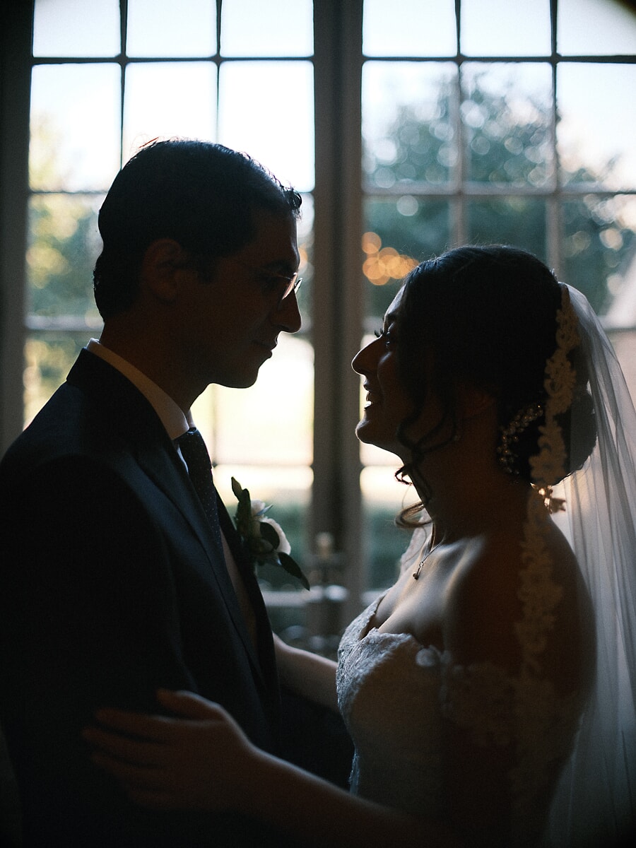 Wedding Bridal SP21 Silhouetted before tall arched windows, the couple stand face to face as light catches the bride's lace veil, a cinematic bridal portrait