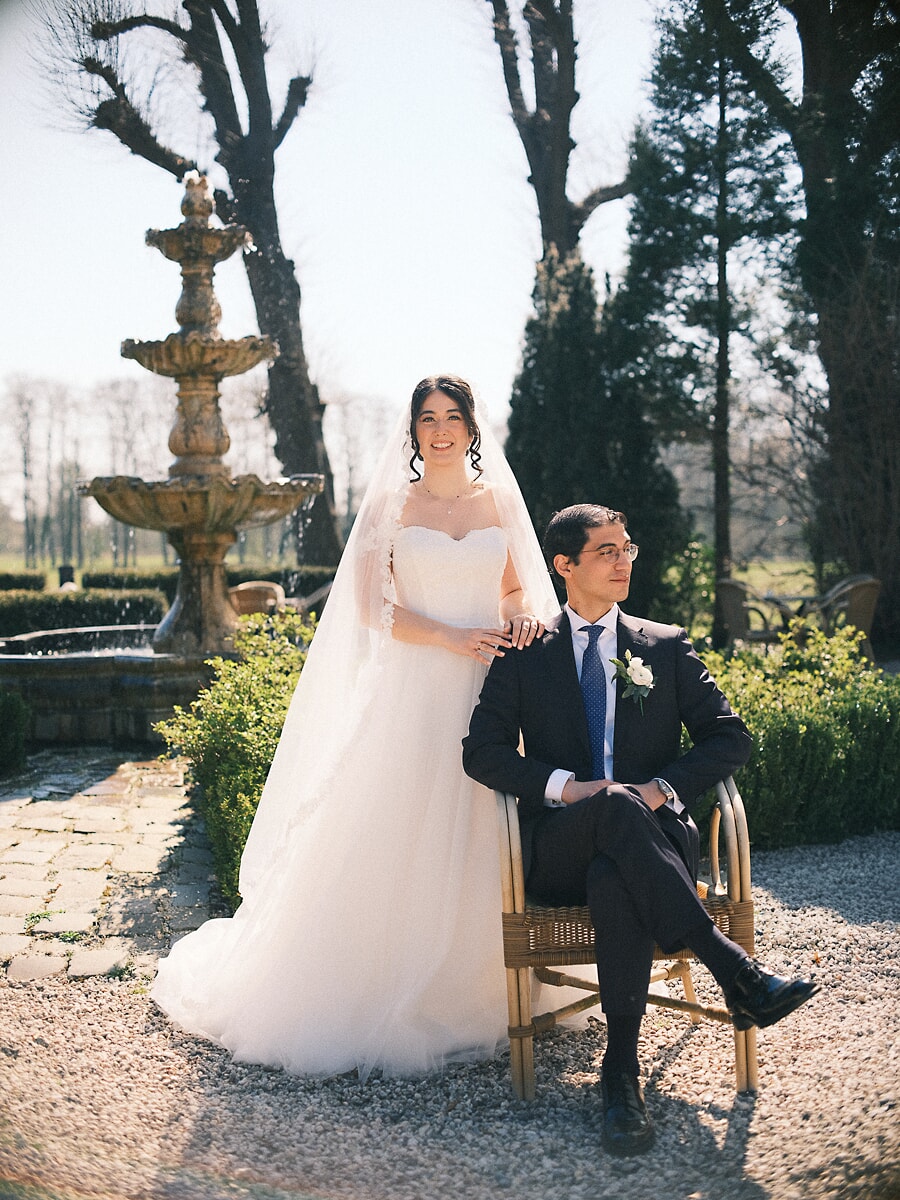 Wedding Bridal SP23 Beside a stone fountain in a bare winter garden, the bride rests a hand on her seated groom's shoulder, an elegant elopement photo