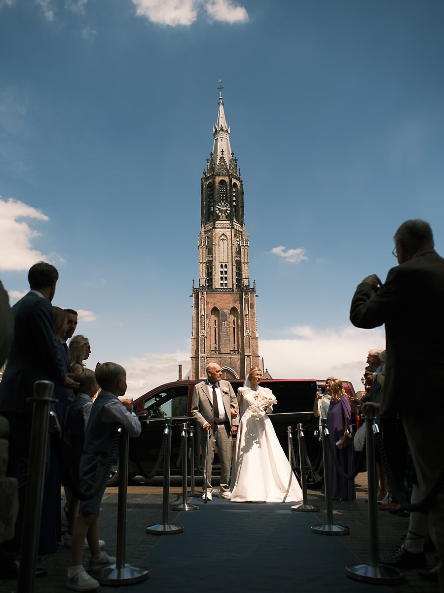 Wedding SP1 A wedding photographer in Amsterdam captures the bride stepping from a dark car onto the blue carpet before the Nieuwe Kerk tower