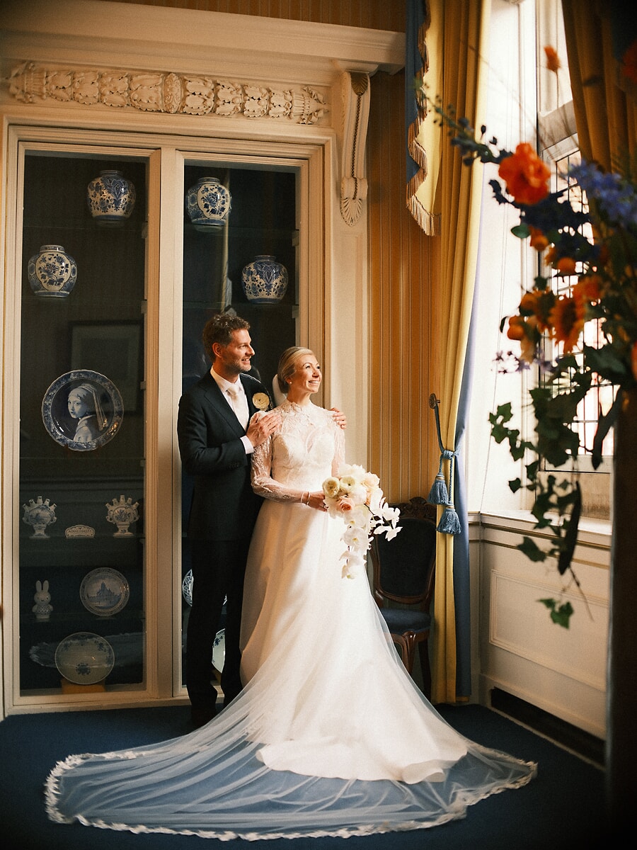 Wedding SP10 Newlyweds pose beside a cabinet of Delft blue porcelain, the bride's veil spread across the floor, an elegant wedding portrait