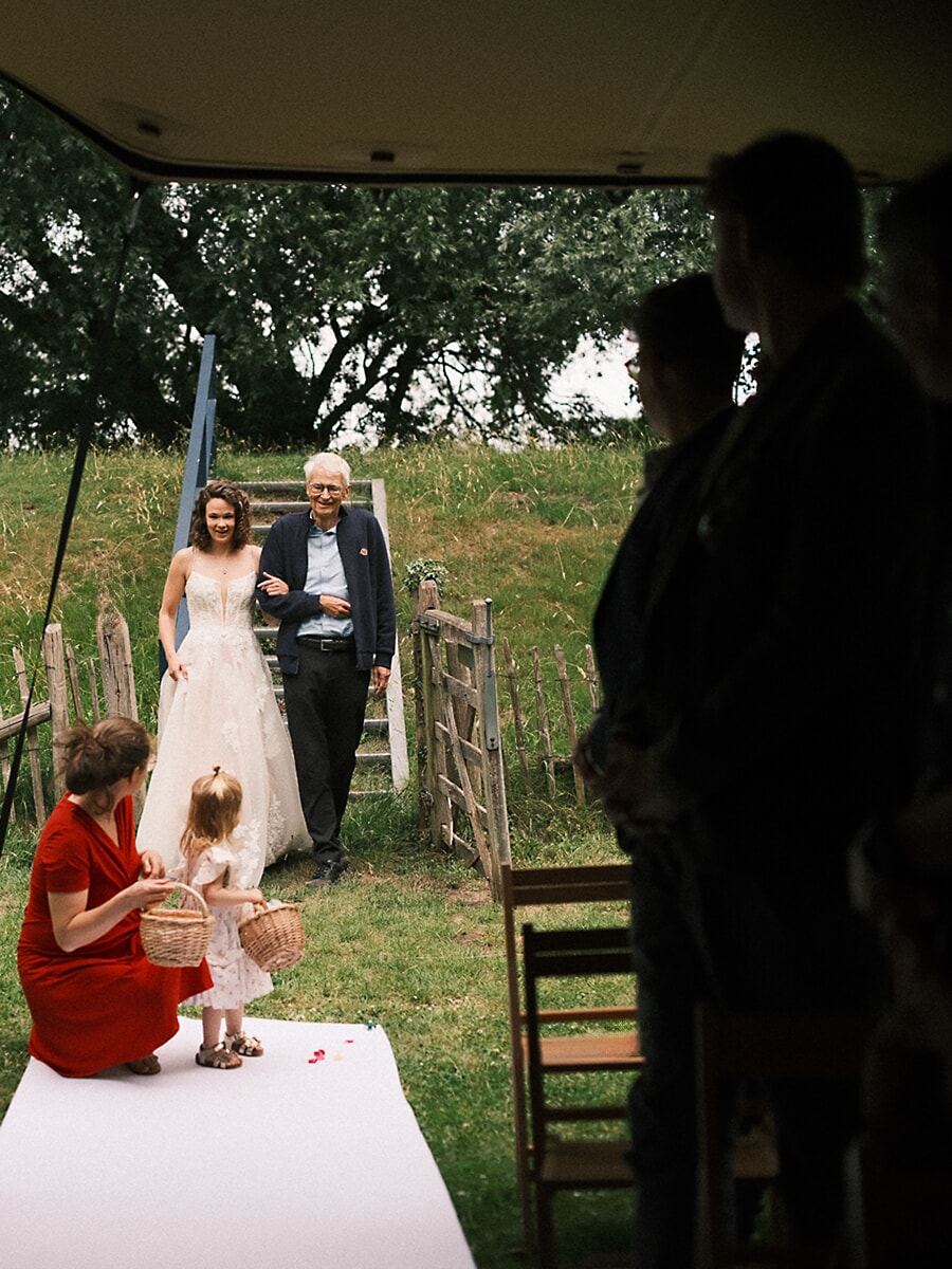 Wedding SP16 Walking the white aisle runner on her father's arm, the bride passes a flower girl filling her basket with petals, a spontaneous ceremony photo