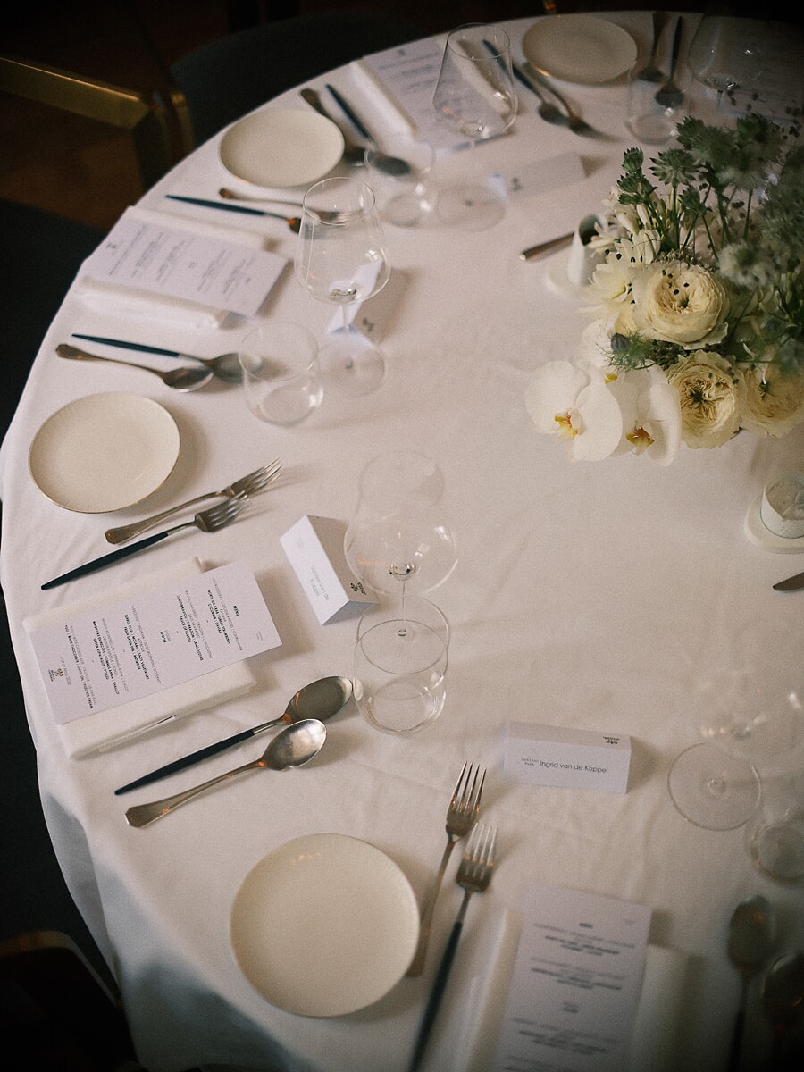 Wedding SP18 Overhead view of a round table in white linen with menu cards, silver cutlery and pale ranunculus, a refined wedding table detail