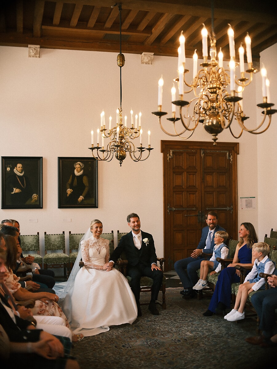 Wedding SP2 Newlyweds sit side by side under brass candle chandeliers as family and children watch the ceremony, a natural marriage photograph