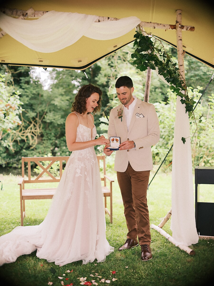 Wedding SP21 Exchanging rings beneath a birch and ivy arch, the couple smile at the open ring box, captured by a wedding photographer in Amsterdam