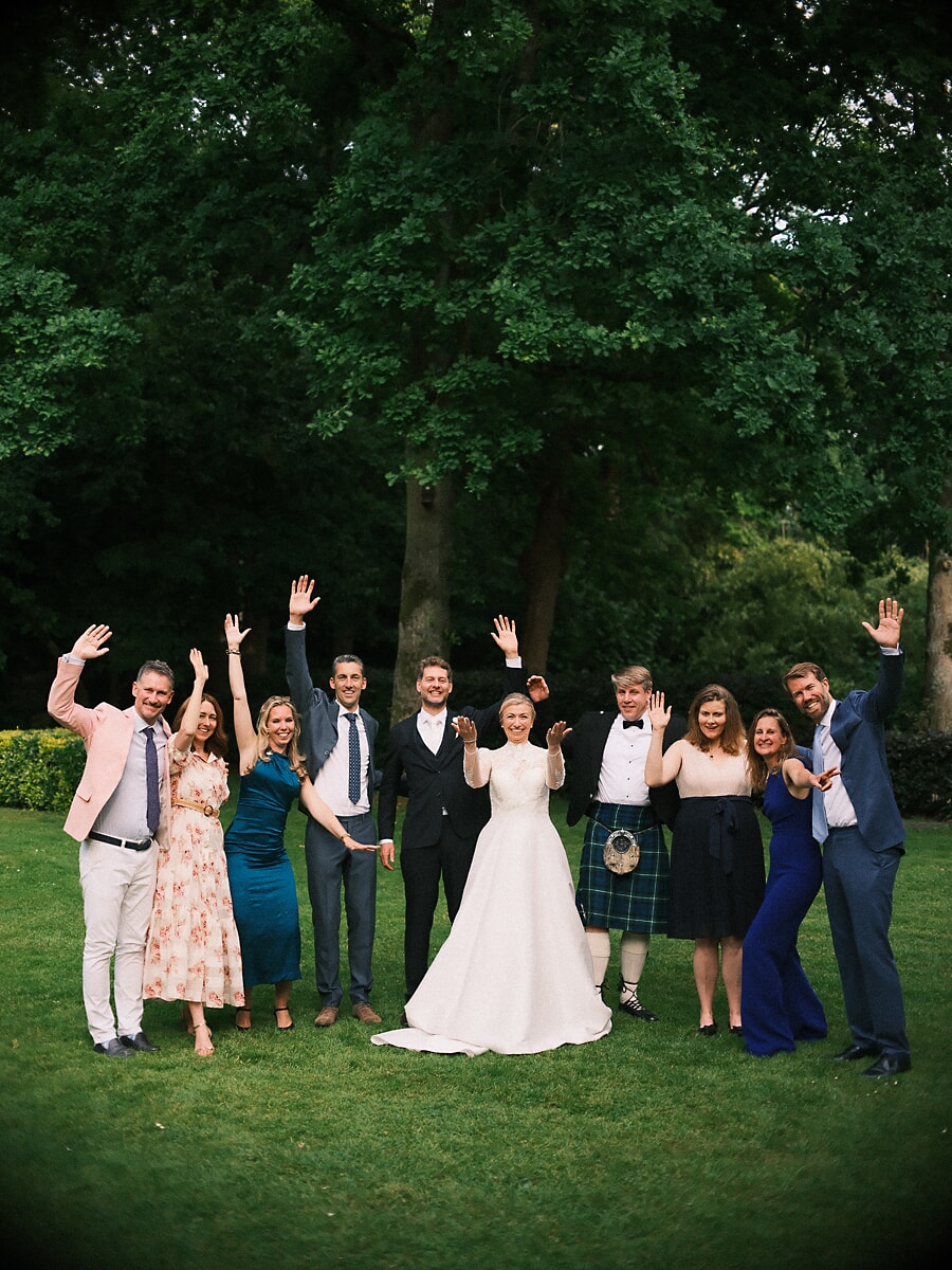 Wedding SP22 Wedding party throw their hands in the air on a green lawn, one guest in a tartan kilt beside the beaming bride, a lively group photo